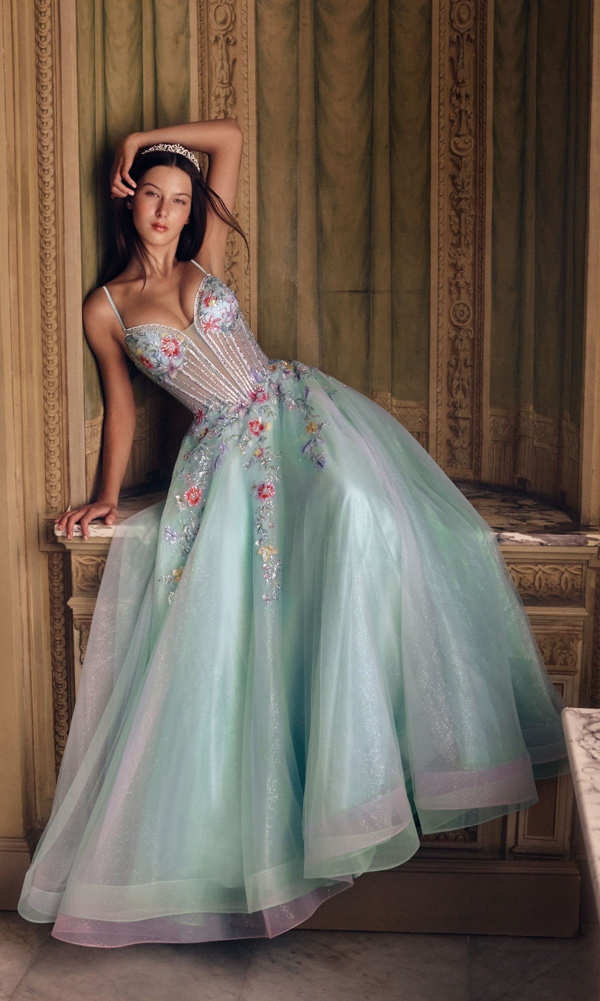 A woman in the Alyce 62068 long prom dress in Sea Glass, featuring a floral corset bodice and sparkly tulle A-line skirt, stands against ornate walls in an elegant room.